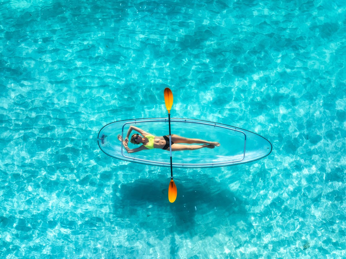 A person lying on a clear transparent canoe in turquoise Caribbean water, captured from a drone above