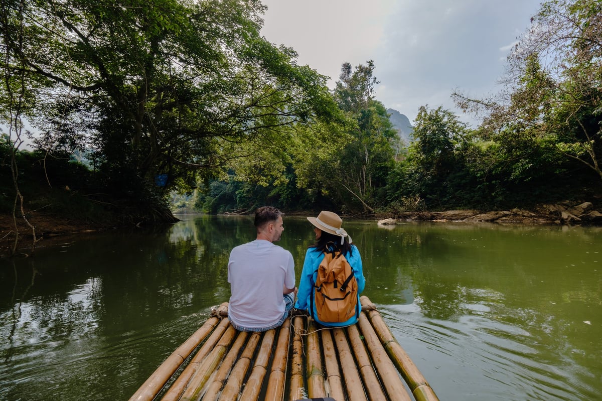 Two travelers floating on a bamboo raft along a peaceful river surrounded by lush greenery