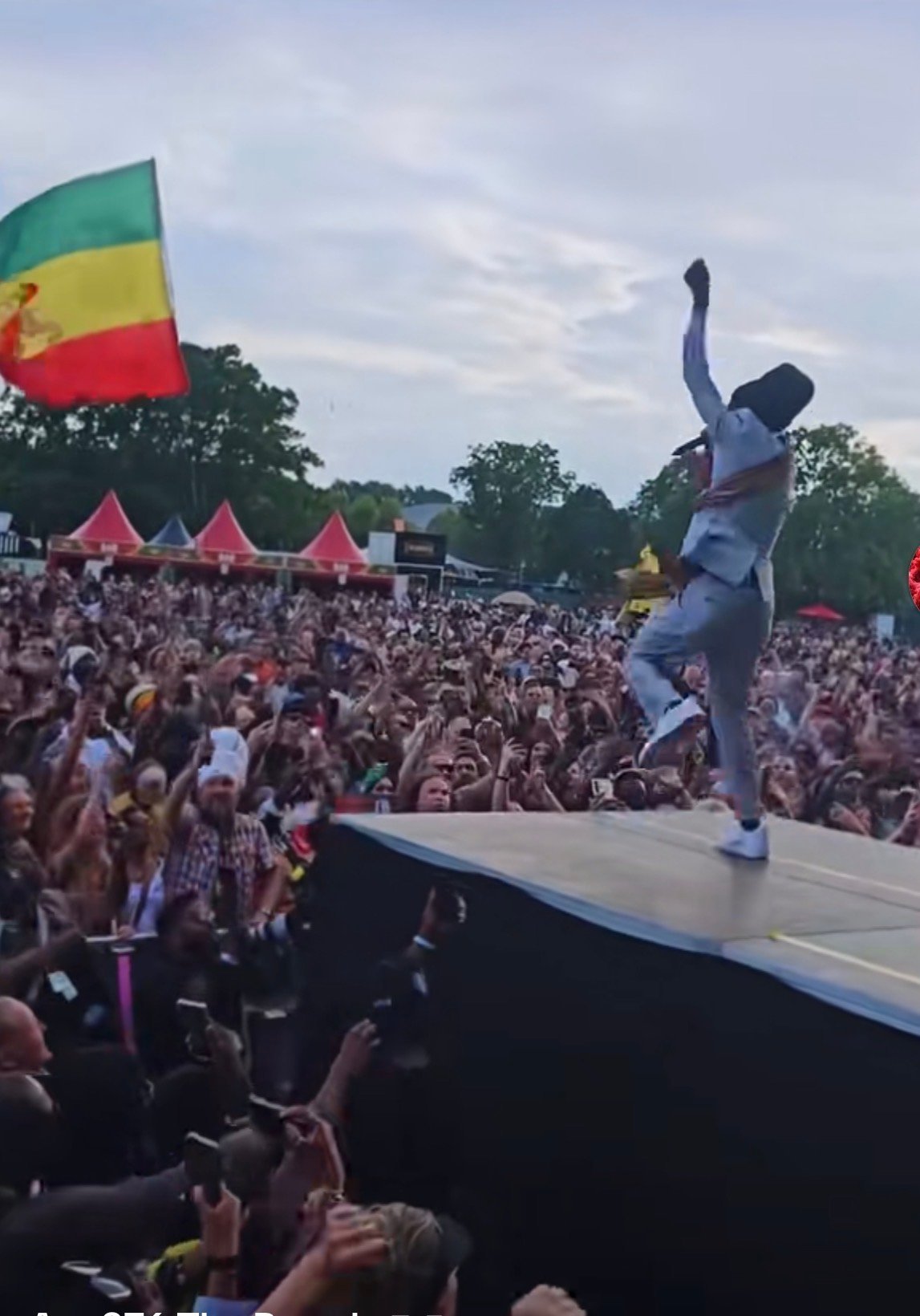 Performer on stage with arm raised at outdoor festival, large crowd visible, Rastafarian flag in background