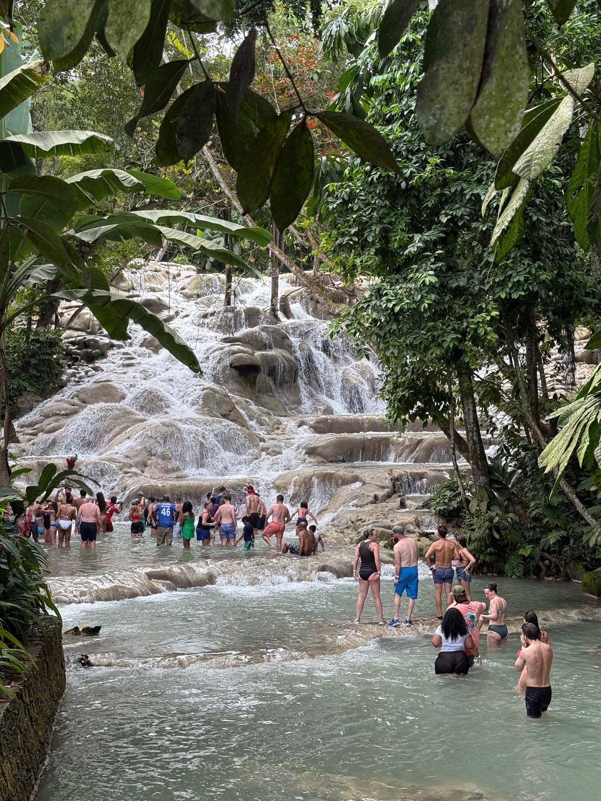 Tourists swimming and wading in turquoise pool beneath multi-tiered waterfall surrounded by lush green jungle vegetation