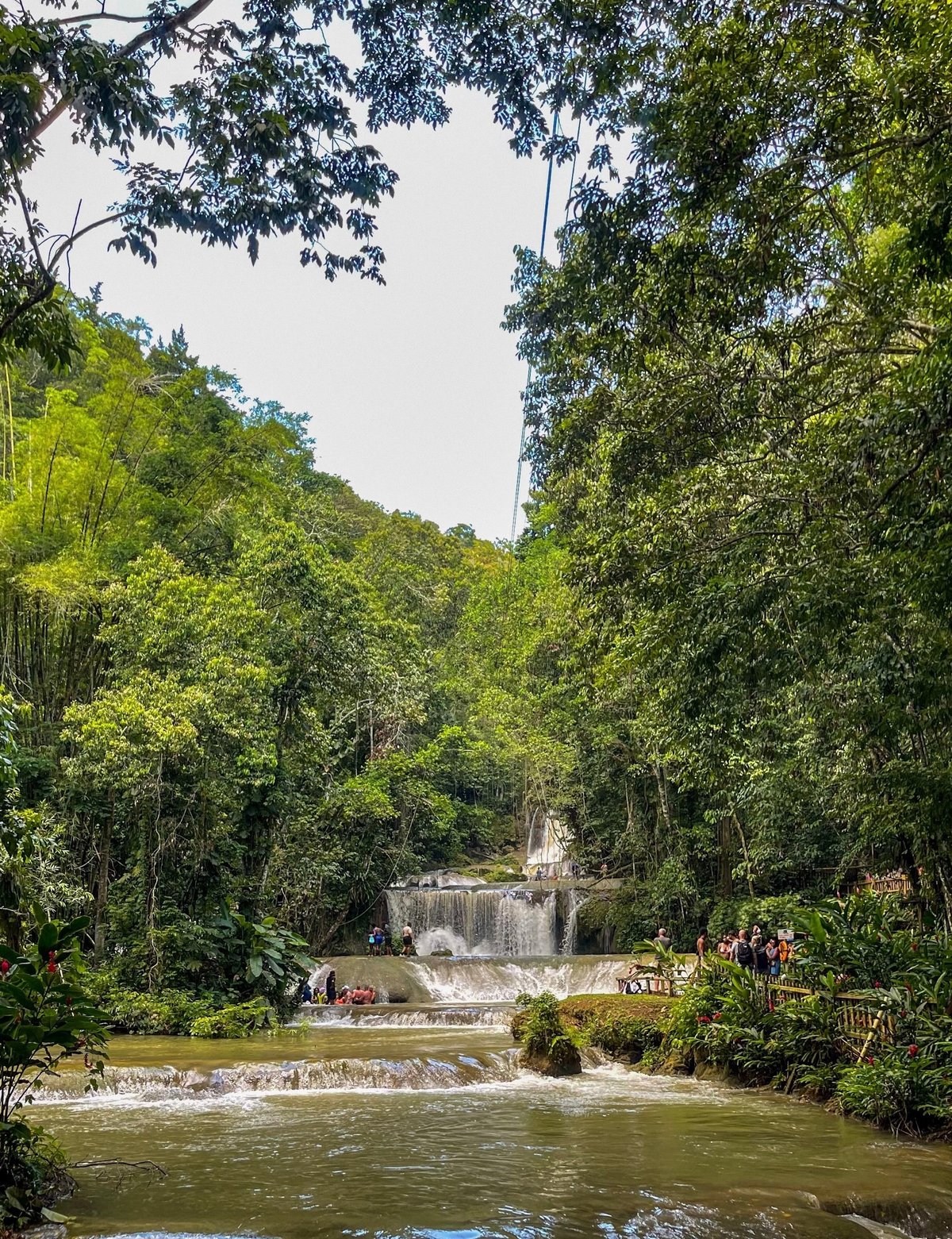 Tiered waterfall cascading through lush green forest with water pools in foreground and visitors on bridge at YS Falls, St Elizabeth