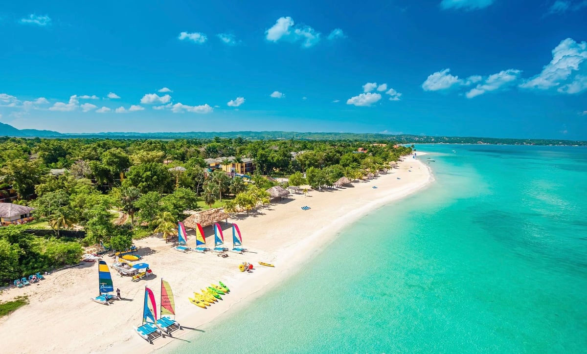 Aerial view of a tropical beach with turquoise water, white sand, colorful sailboats, and lush green vegetation along the shoreline at Seven Mile Beach, Negril