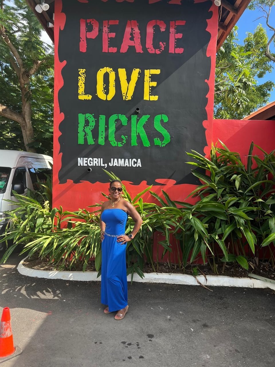 Woman in blue dress posing in front of Peace Love Ricks sign in Negril Jamaica with tropical plants