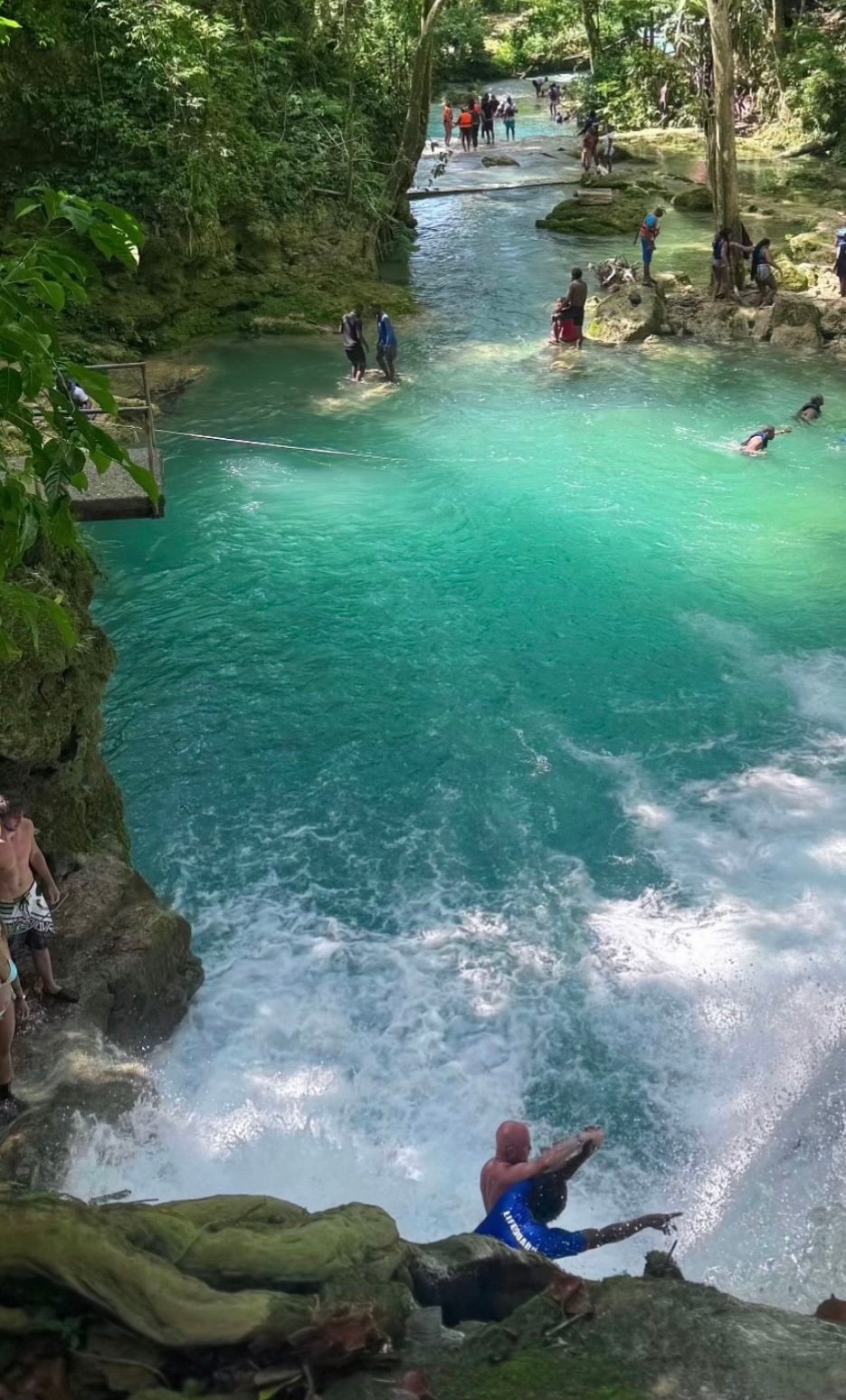 Person diving into turquoise lagoon surrounded by lush forest, with swimmers and onlookers enjoying the natural pool