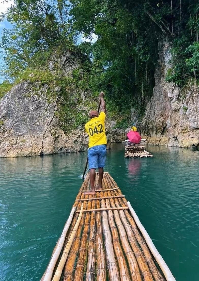 Person in yellow shirt poling a bamboo raft on turquoise water with limestone cliffs and another raft visible in background