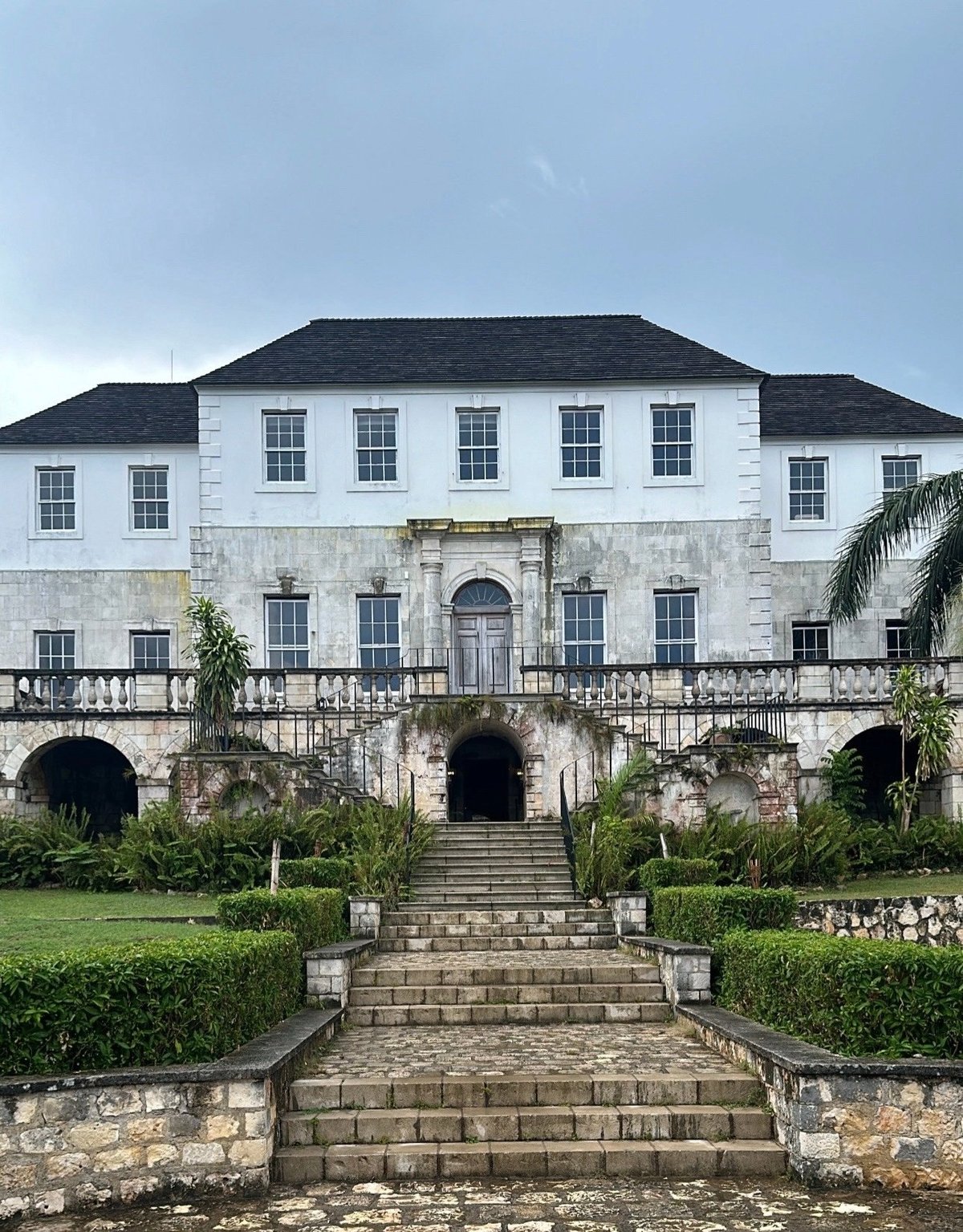 Historic white and gray colonial mansion with dark roof, arched stone entrances, manicured gardens, and palm trees on the grounds at Rose Hall, Montego Bay