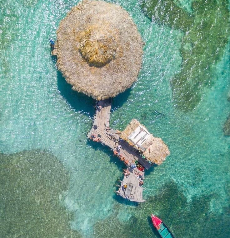 Aerial view of Floyd's Pelican Bar, a thatched-roof wooden bar built on stilts over clear turquoise water, with a small dock and a red boat moored alongside in St Elizabeth